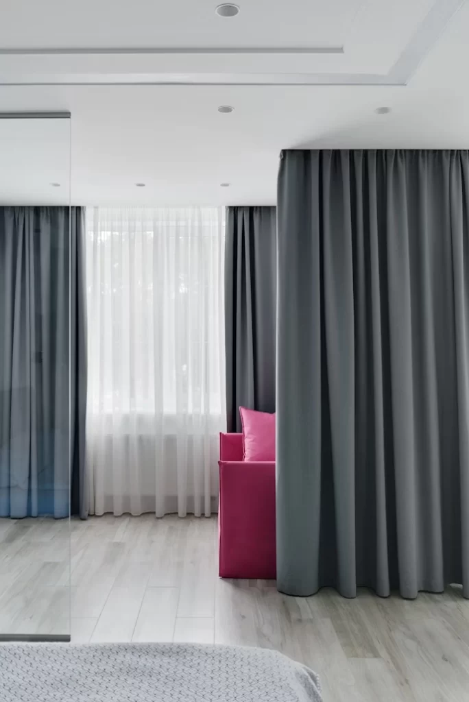 A modern room with light wood floors and white walls. Two large, dark gray blackout curtains hang on the right, partially concealing a bright pink sofa