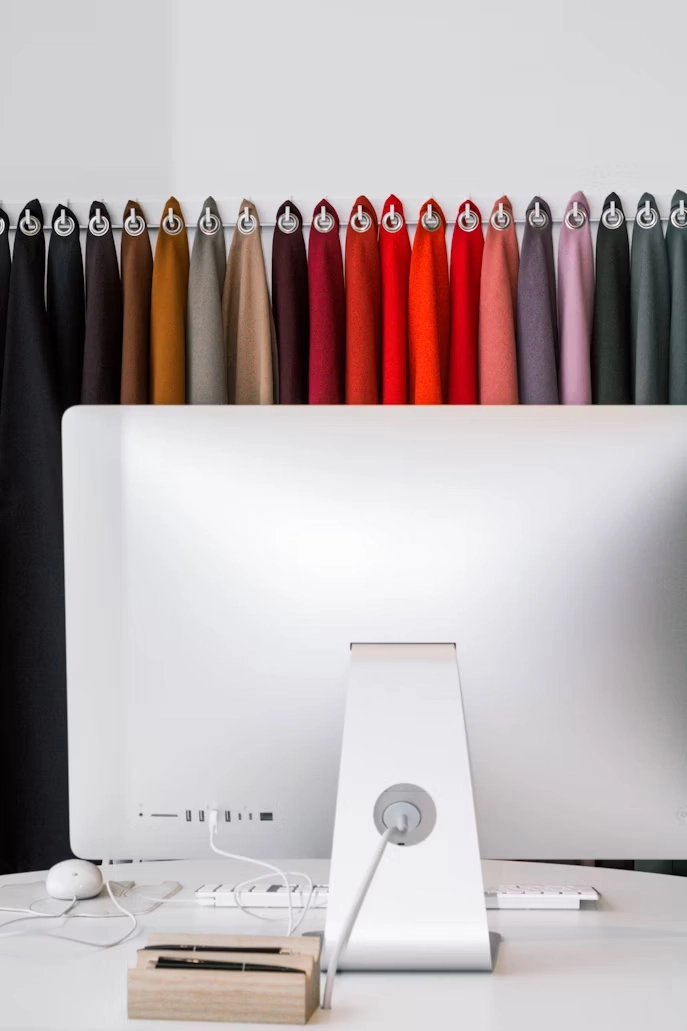 A variety of fabric swatches for blackout curtains, in various colors, are hanging above a computer monitor on a desk