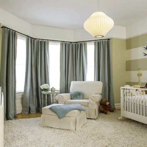 A peaceful, sunlit baby room featuring sage green nurseries blackout curtains installed on a curved black curtain rod along a bay window.