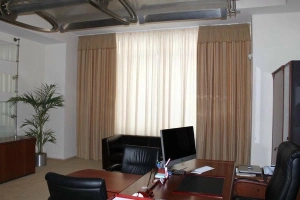 An office space featuring home office blackout curtains in a neutral tan color, which are partially drawn to filter the light from a large window.
