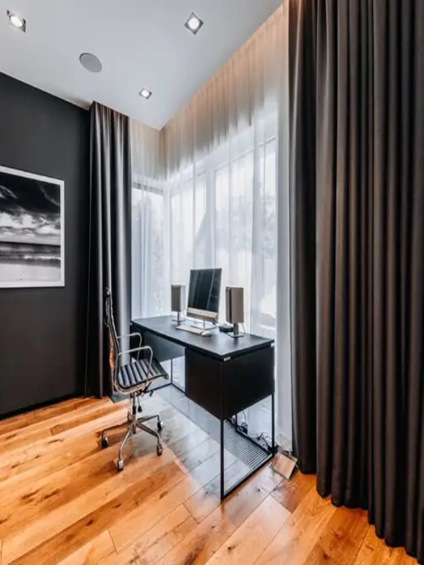 A modern home office featuring floor-to-ceiling charcoal grey blackout curtains that frame a large window with sheer white liners.